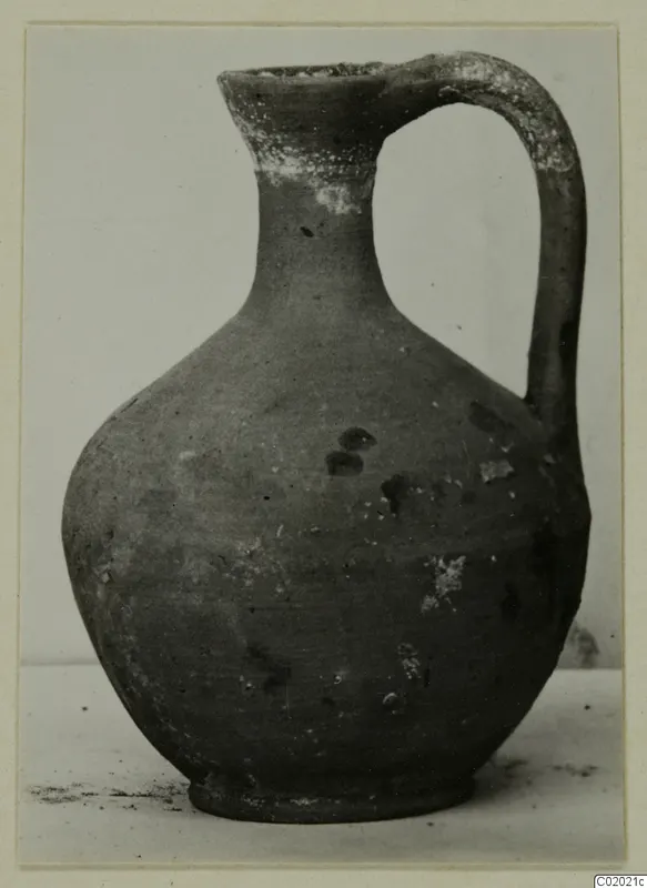 Plain pottery jug with a high loop handle and a rounded body, photographed on a pale surface.