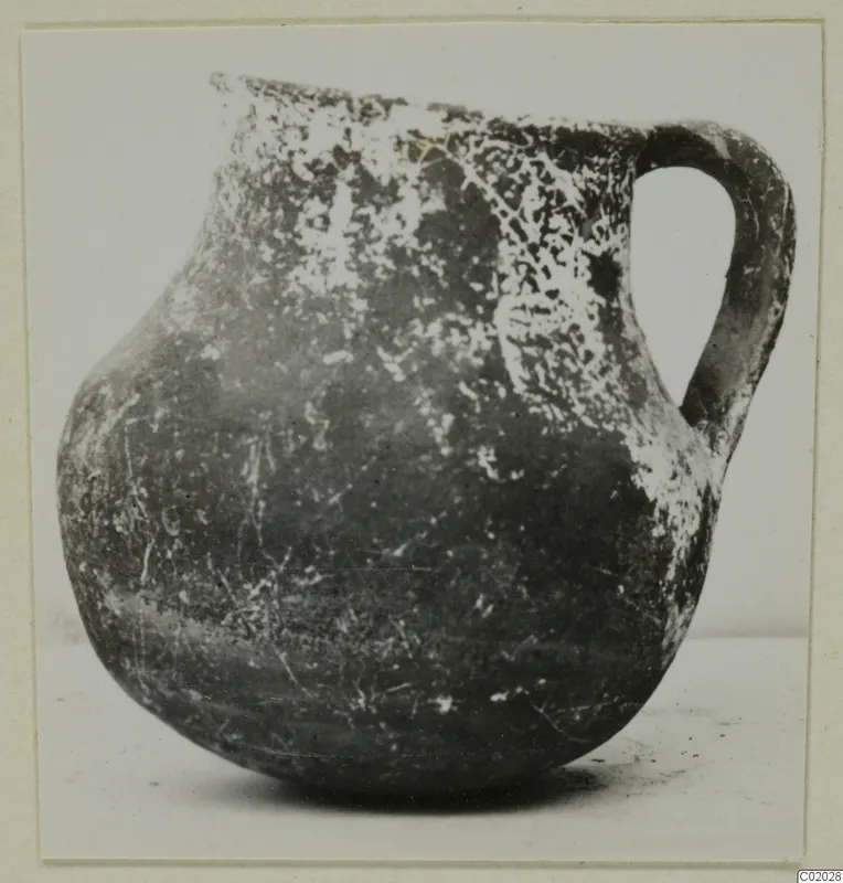 Coarse pottery jug with a single loop handle, dark surface with pale encrusted patches