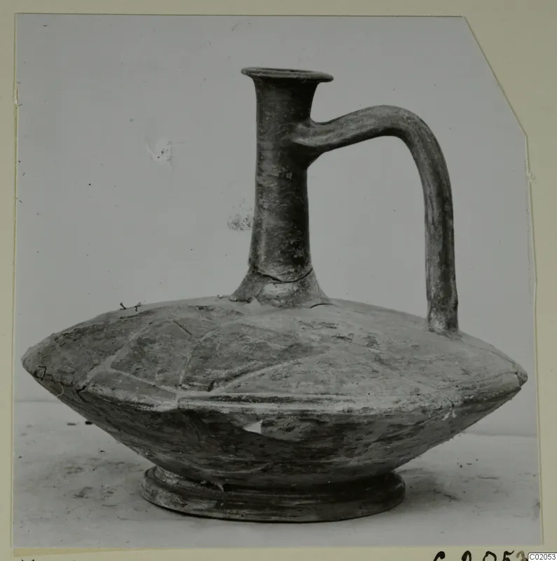 Pottery jug with a tall flared neck, a single loop handle and a flattened, ribbed body on a low foot.