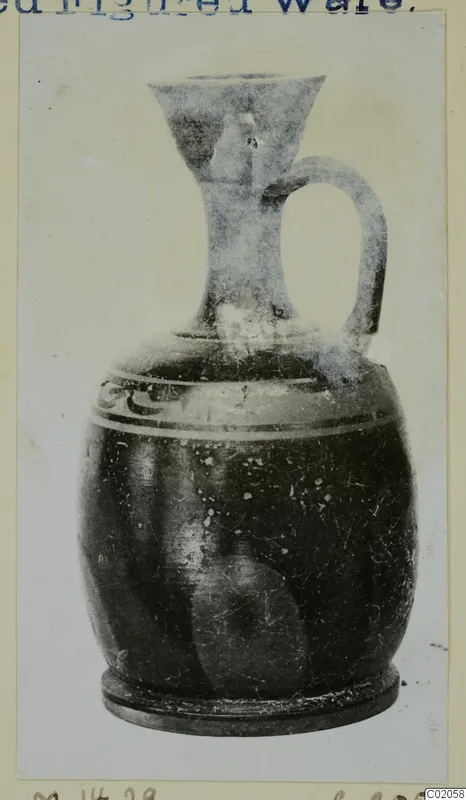 Dark jug with a funnel-shaped mouth, strap handle and pale bands around the body, shown on a mounted print.