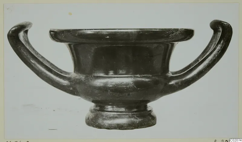 Black glazed kantharos with two curving handles, photographed in side view against a pale background