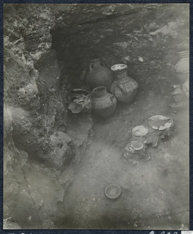 Tomb 84 at Marion: pottery vessels and broken sherds on the floor of a rock-cut tomb chamber during excavation.