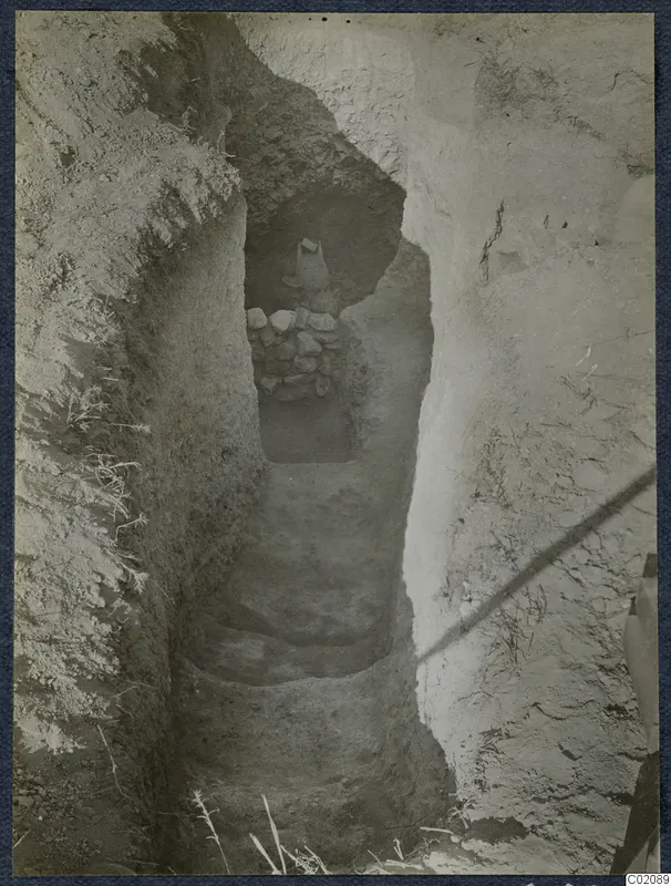 Steps leading down a narrow rock-cut passage at Marion to a tomb opening blocked by stones, with a vessel on top.