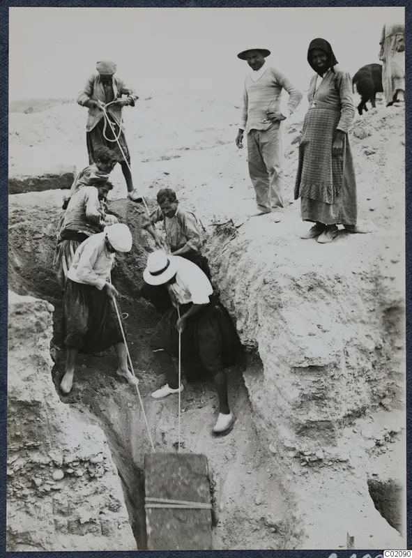 Workers haul a wooden box out of a narrow excavation trench on ropes, with two onlookers standing at the edge