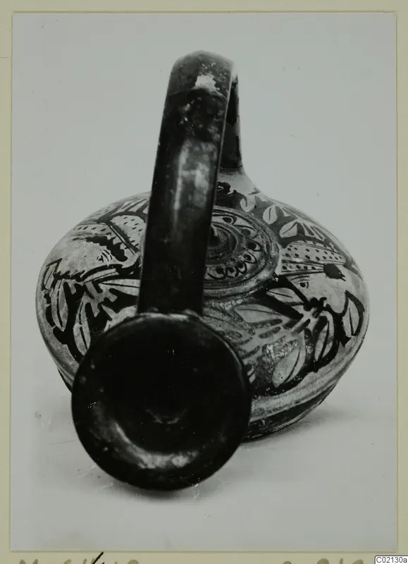 Painted pottery vessel with a loop handle and dark leaf and scroll decoration, mouth turned toward the camera.