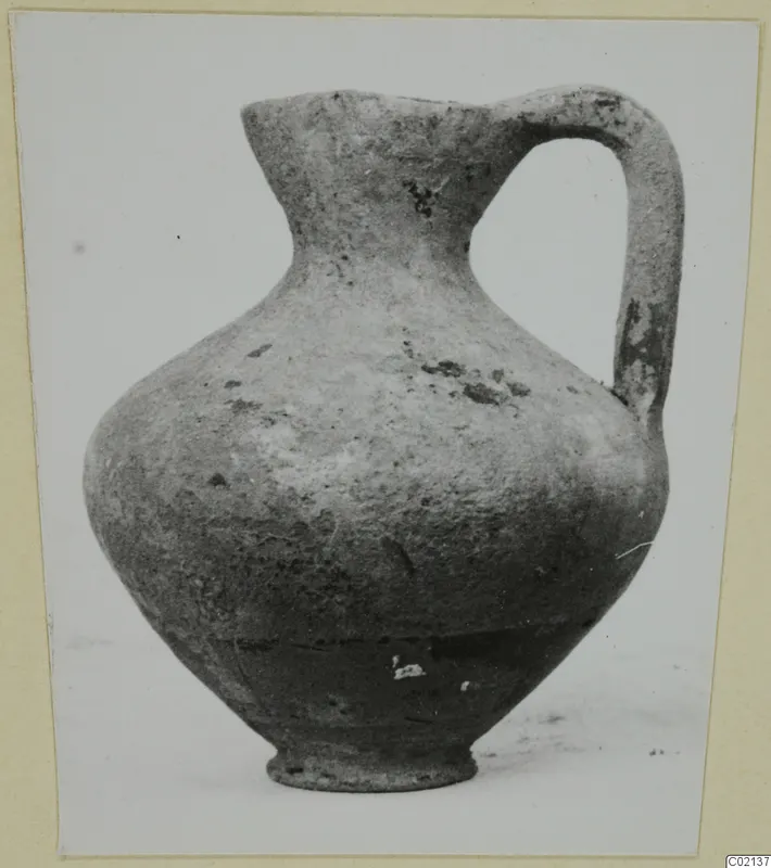 Pottery juglet with a single strap handle and a flaring neck, shown on a pale surface
