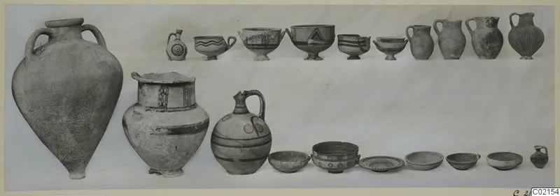 Group of pottery vessels, jugs, bowls and a two-handled amphora, arranged in two rows against a pale background.