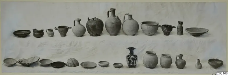 Two rows of pottery vessels on a white cloth, including jugs, cups, jars and shallow bowls