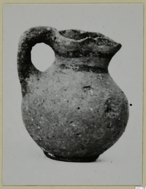 Small juglet with a loop handle and rounded body, photographed on a grey card mount