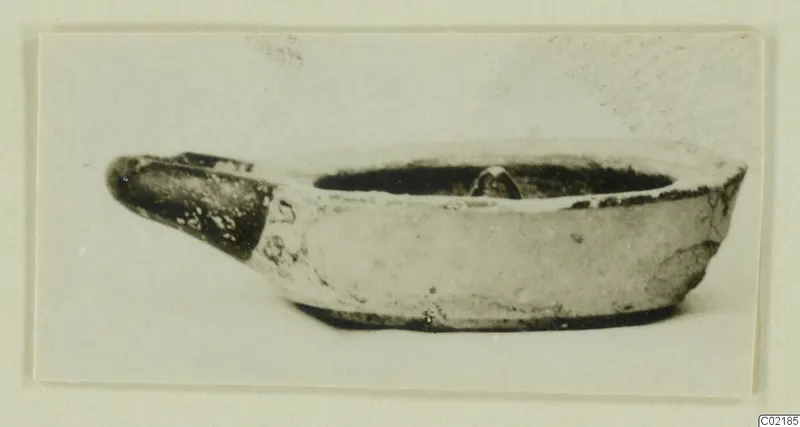 Small terracotta lamp with a pinched spout, dark glaze on the spout and rim, shown on a mounted card.