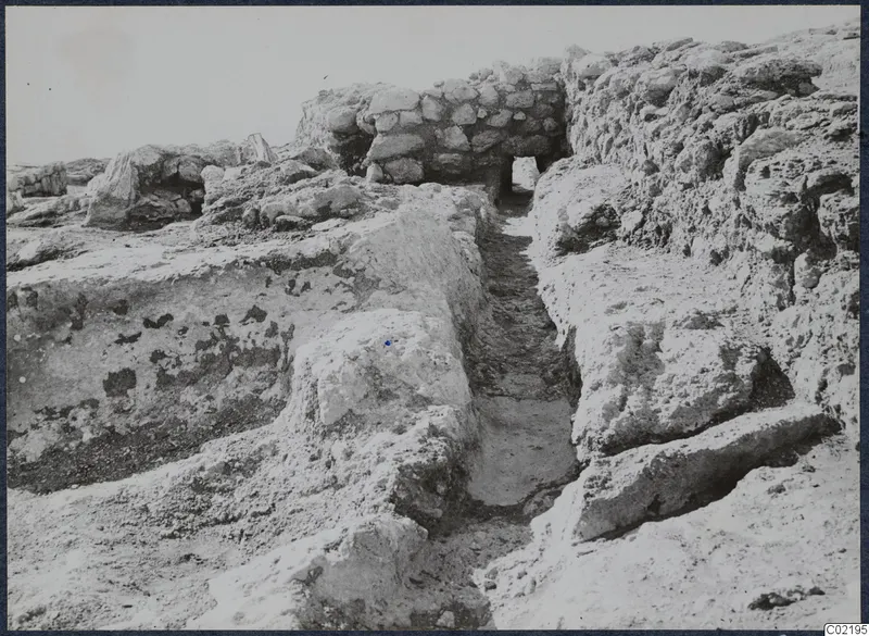 Excavated walls and loose stones at the palace at Vouni, with a narrow cut passage running between them.