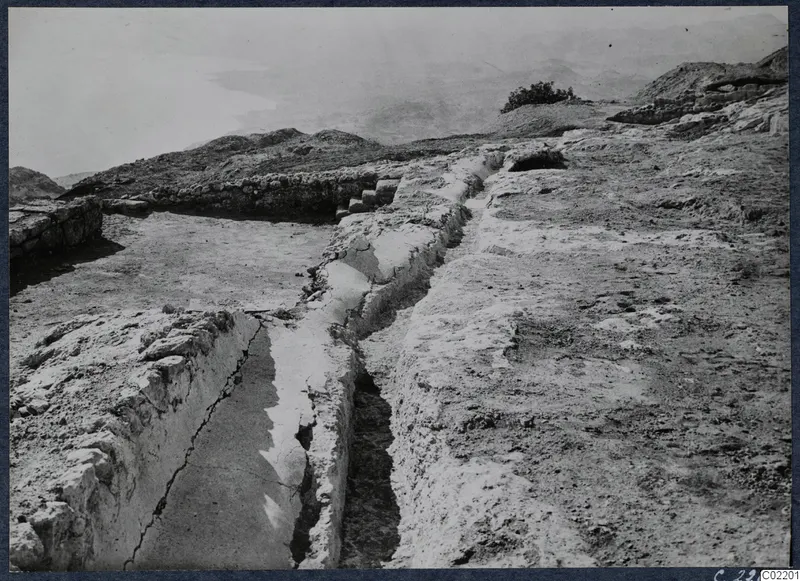 Excavated rock-cut channel and low walls at the palace at Vouni, with hills and a pale open view beyond.