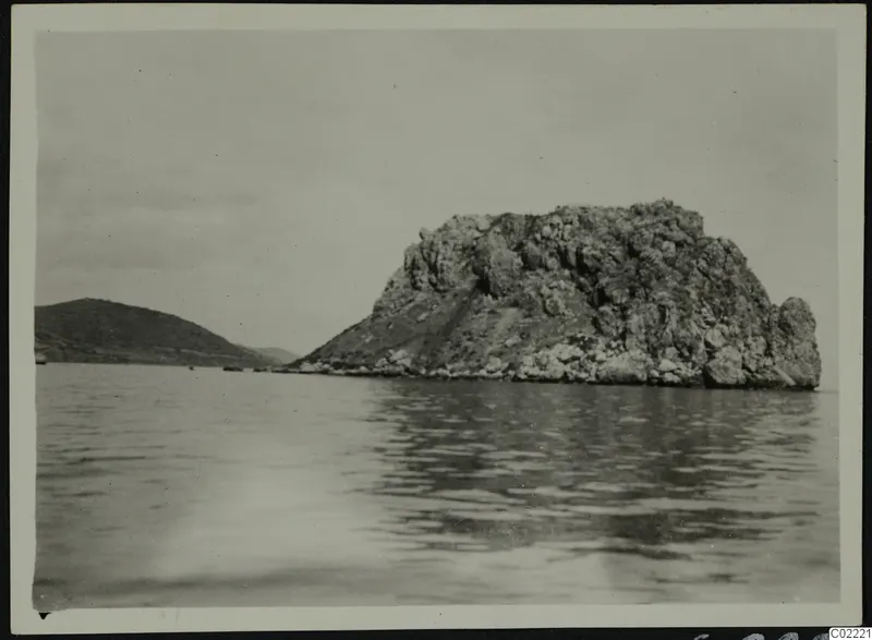 Rocky islet rising from the sea at Petra tou Limniti, with a low hill on the left.