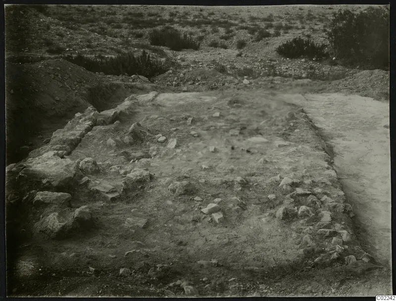 Excavated area at Ayios Iakovos with exposed stone walls, scattered stones and low scrub beyond.