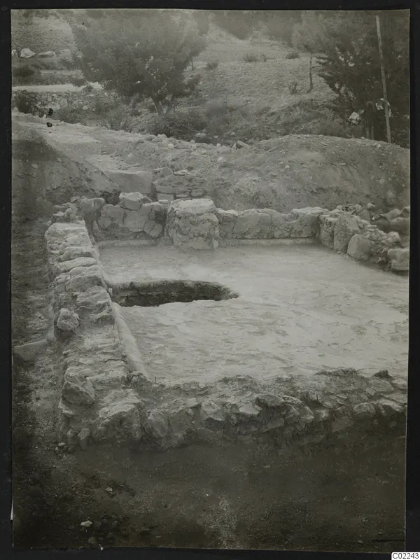 Excavated area at Ayios Iakovos with a low stone wall enclosing a pale floor and a round pit