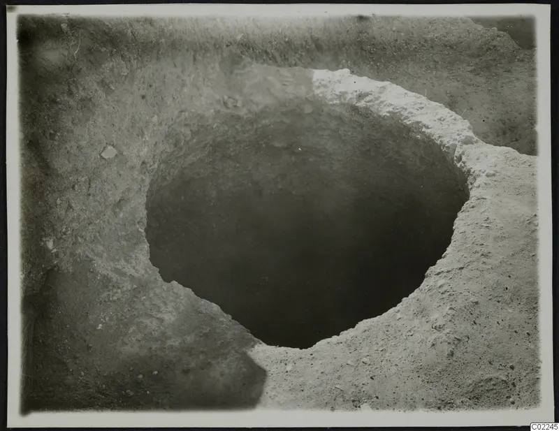 Round pit cut into excavated soil at Ayios Iakovos, with small stones scattered in the surrounding ground.