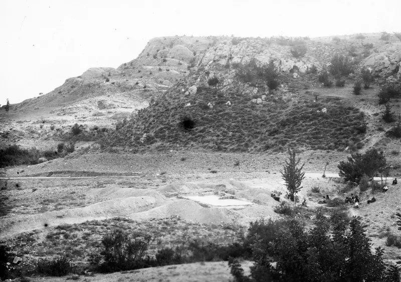 Excavation area at Ayios Iakovos below a rocky hillside, with spoil heaps, a pale cut in the ground and people seated under trees