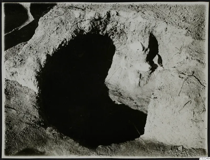 Dark round opening cut into pale earth at Ayios Iakovos, with white stones set in the soil beside it.