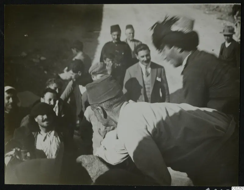 Men gathered on a dusty road, one bending over a pale rounded object, others standing and seated around him in caps and fezzes