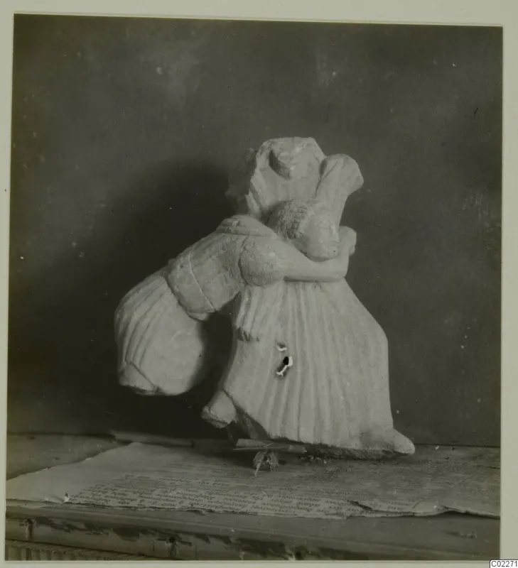 Carved limestone figure in a long pleated garment, one arm around a bulky rounded form, head and upper body broken