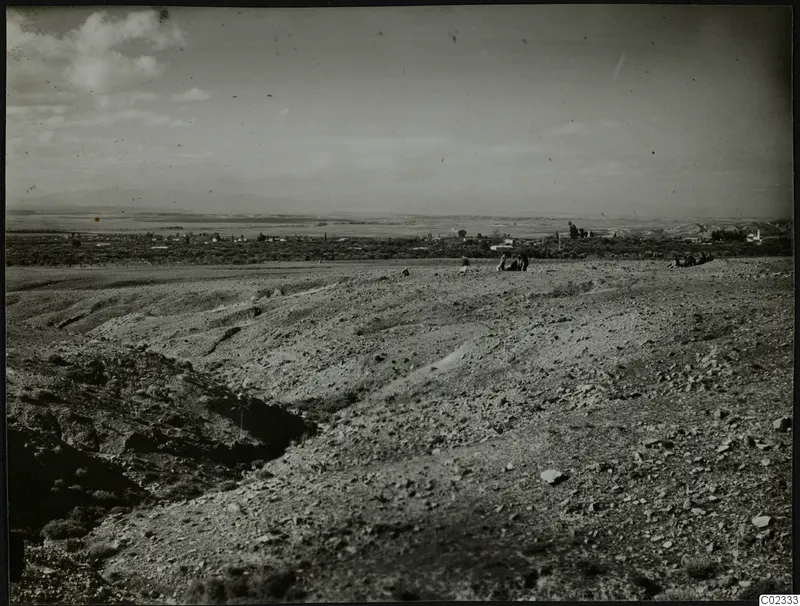Stony hillside at Kythrea with a few people, a gully on the left and a village on the plain below distant hills