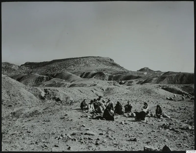 People seated on stony ground below eroded hills at Kythrea, with a flock of sheep or goats at the right edge.