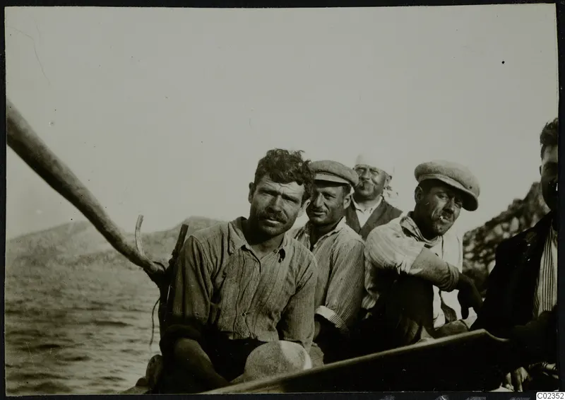 Petra tou Limniti: five men seated in a wooden boat on the water, with hills and a rocky shore behind them.