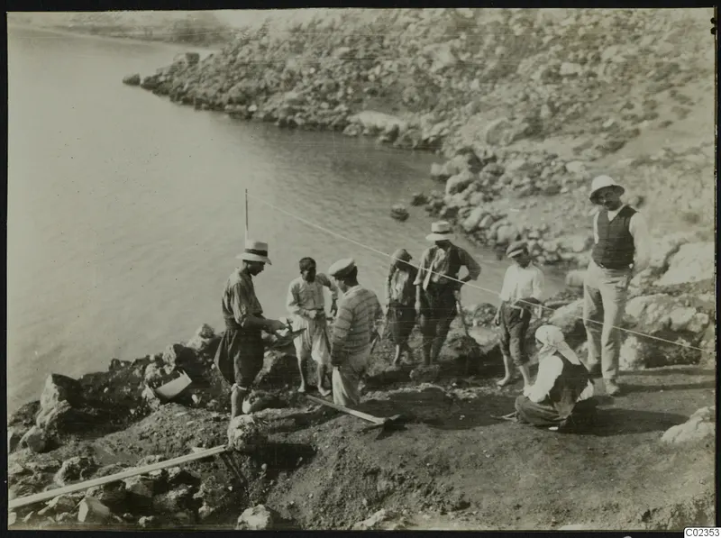 Workers and a woman in headscarf at an excavation on a rocky shoreline at Petra tou Limniti, with a cord stretched across the dig.
