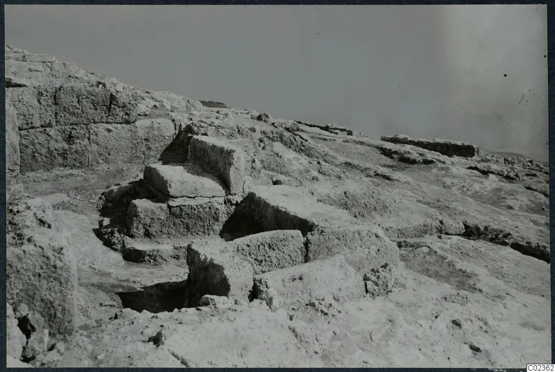 Excavated stone walls and blocks on a rocky slope at Vouni, with a dark cut opening in the foreground.