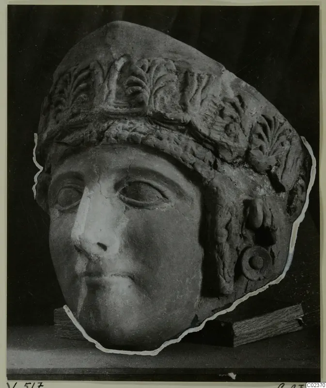 Sculpted head of a figure wearing a tall headdress carved with leaf motifs, shown on wooden blocks against a dark backdrop.