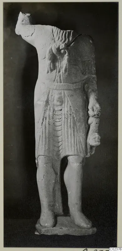Headless limestone statue of a standing figure in a short pleated garment, on a base