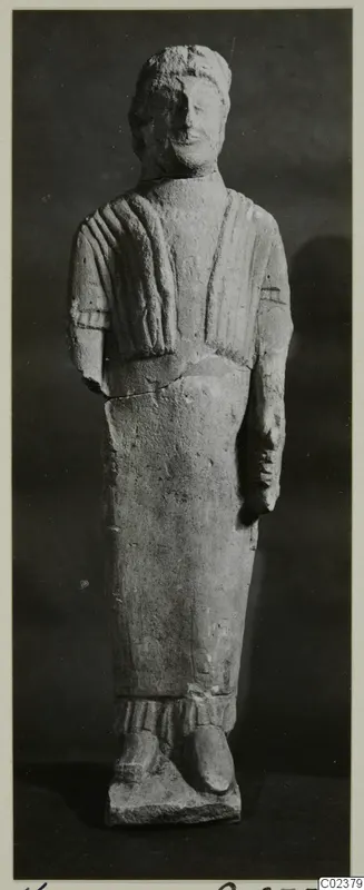 Limestone statue of a standing figure in a draped cloak, arms at its sides, shown against a dark backdrop