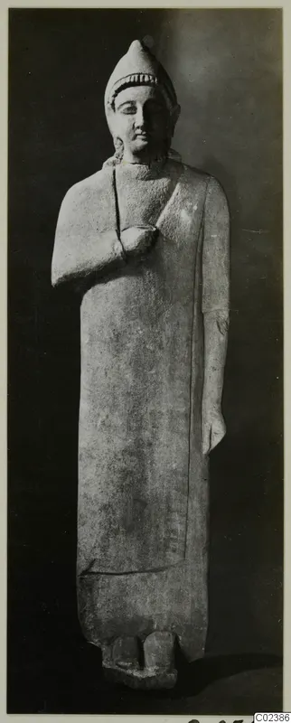 Limestone statue from Kition of a standing figure in a tall cap and long robe, one hand raised to the chest.