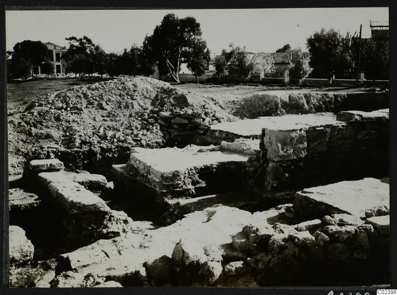 Excavated stone walls and large cut blocks at Bamboula, Kition, with a spoil heap behind and trees beyond.