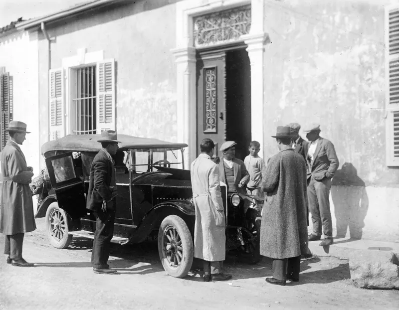 Men in hats, caps and long coats stand around a black open car outside a plastered building with a carved doorway.