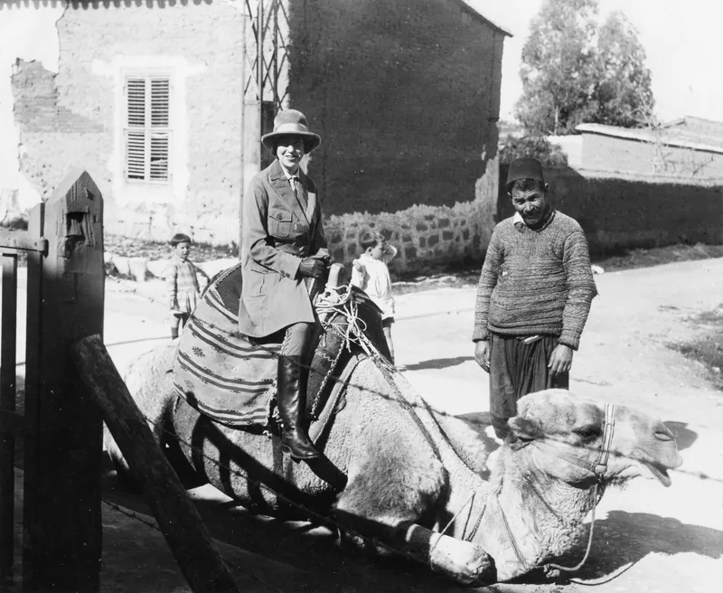 Vivi Gjerstad riding a camel in a Nicosia street, with a man and two boys beside it