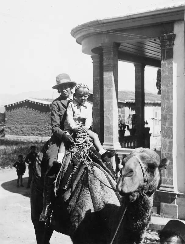 Vivi and Stefan Gjerstad riding a camel past a stone-columned building in Nicosia
