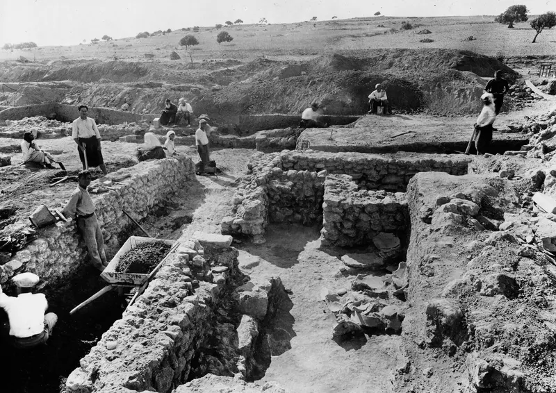 Excavation of a stone house at Ayia Irini, with workers, a wheelbarrow and broken pottery on the floor.