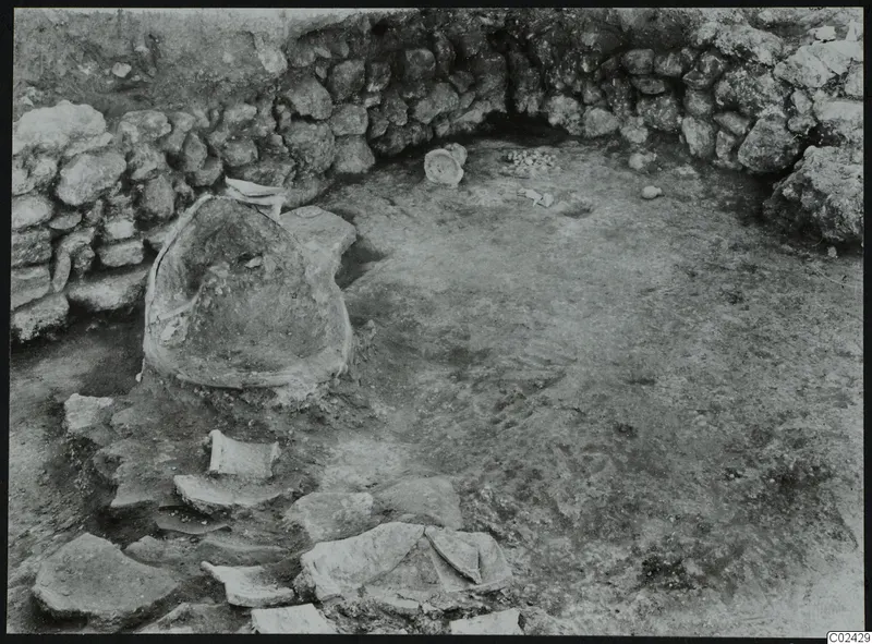 Excavation at Ayia Irini showing a curved stone wall, a large partly buried vessel and scattered sherds.