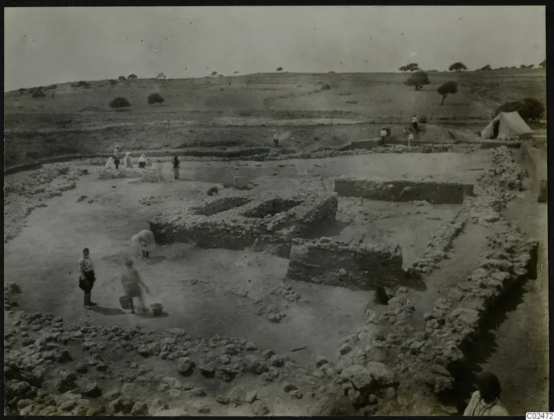 Excavation at Ayia Irini with low stone walls, people carrying buckets across the dug area and a white tent on the right