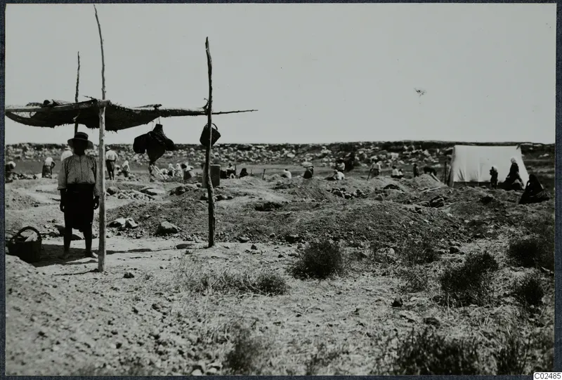 Excavation at Enkomi with workers among spoil heaps, a white tent, and a shaded shelter of poles and matting.