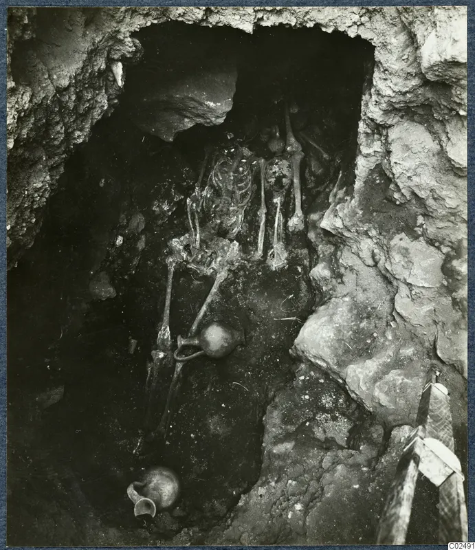 Tomb 14 at Enkomi: skeleton lying in a rock-cut tomb chamber during excavation, with a jug beside it.
