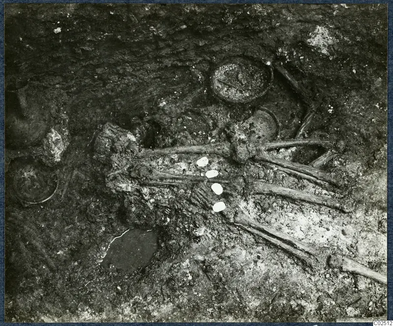 Tomb chamber at Enkomi during excavation, with a skeleton lying in dark soil among pottery vessels