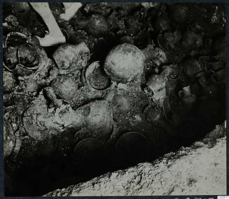 Tomb 13 at Enkomi: a dense cluster of pottery vessels in the tomb chamber during excavation, with a person's legs at the top edge.