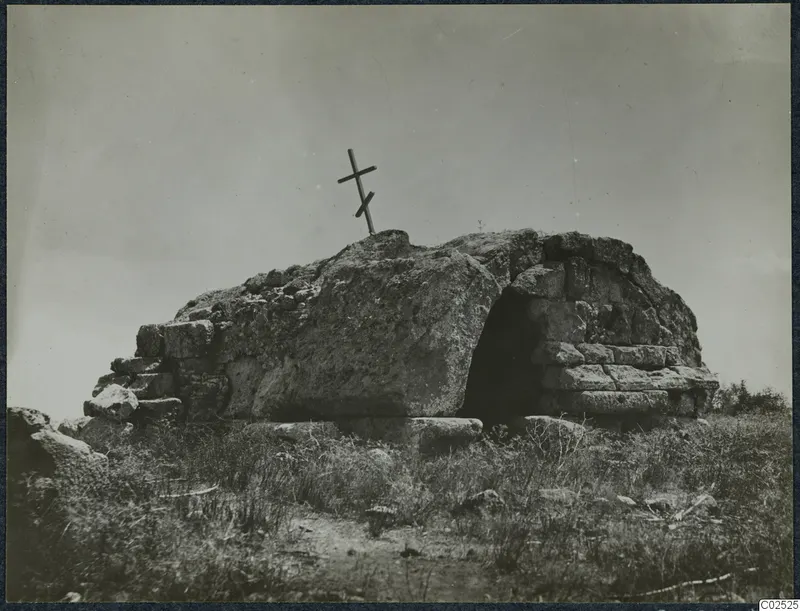 Built tomb of large stone blocks with a dark entrance opening, a wooden cross on top, grass in the foreground