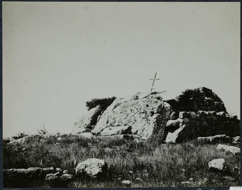 Built stone structure with a wooden cross on top, set among grass and boulders