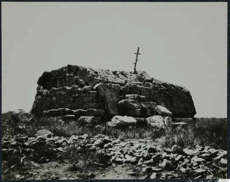 Built stone tomb with a wooden cross on top, surrounded by grass and scattered stones.