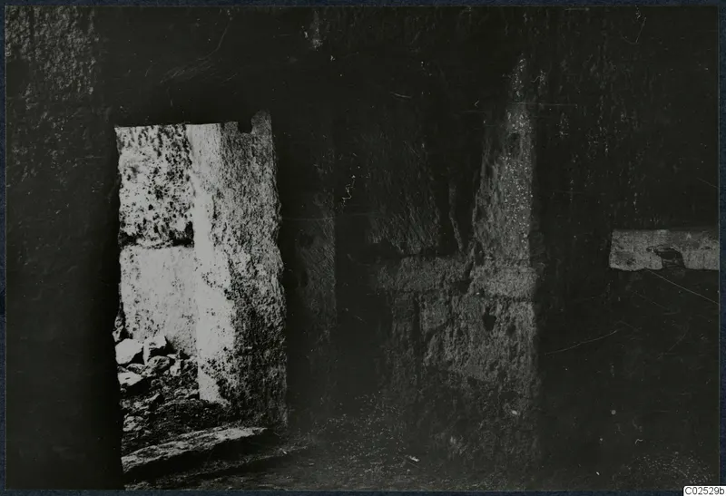 Interior of a built tomb chamber during excavation, with stone walls, a bright doorway on the left and rubble on the floor