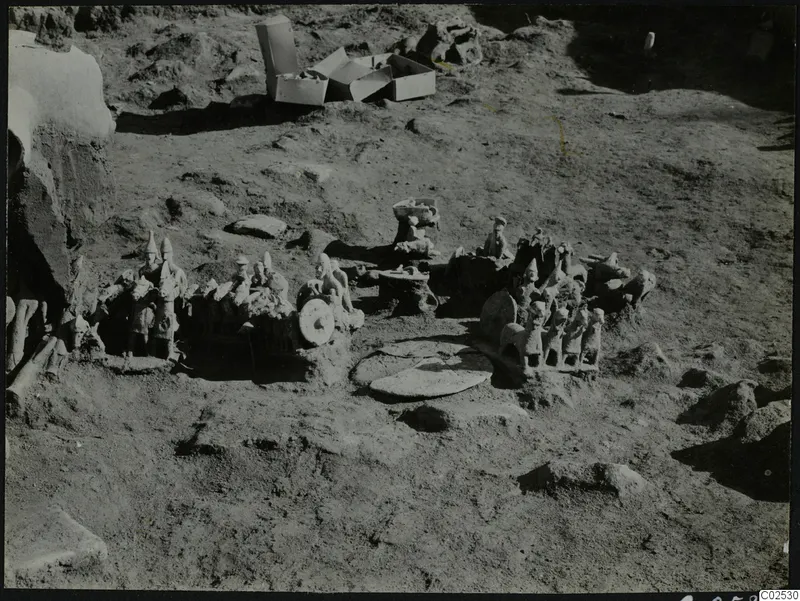 Terracotta figurines and carts lying in excavated ground at Ayia Irini, with cardboard boxes behind.