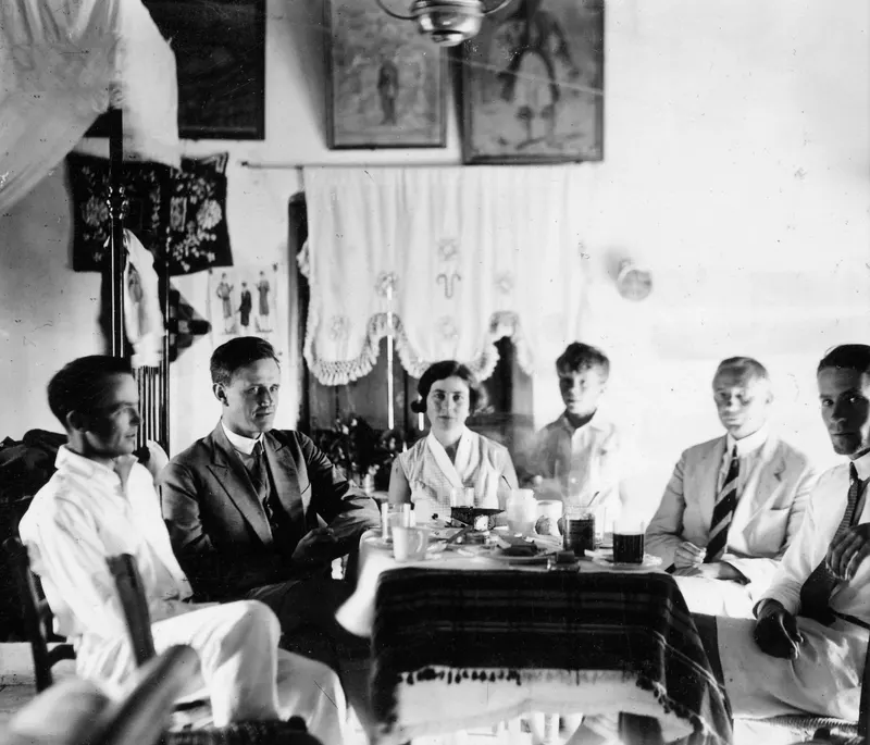 Six people seated at a table in a room at Ayia Irini, with framed paintings and a curtain on the wall behind them.